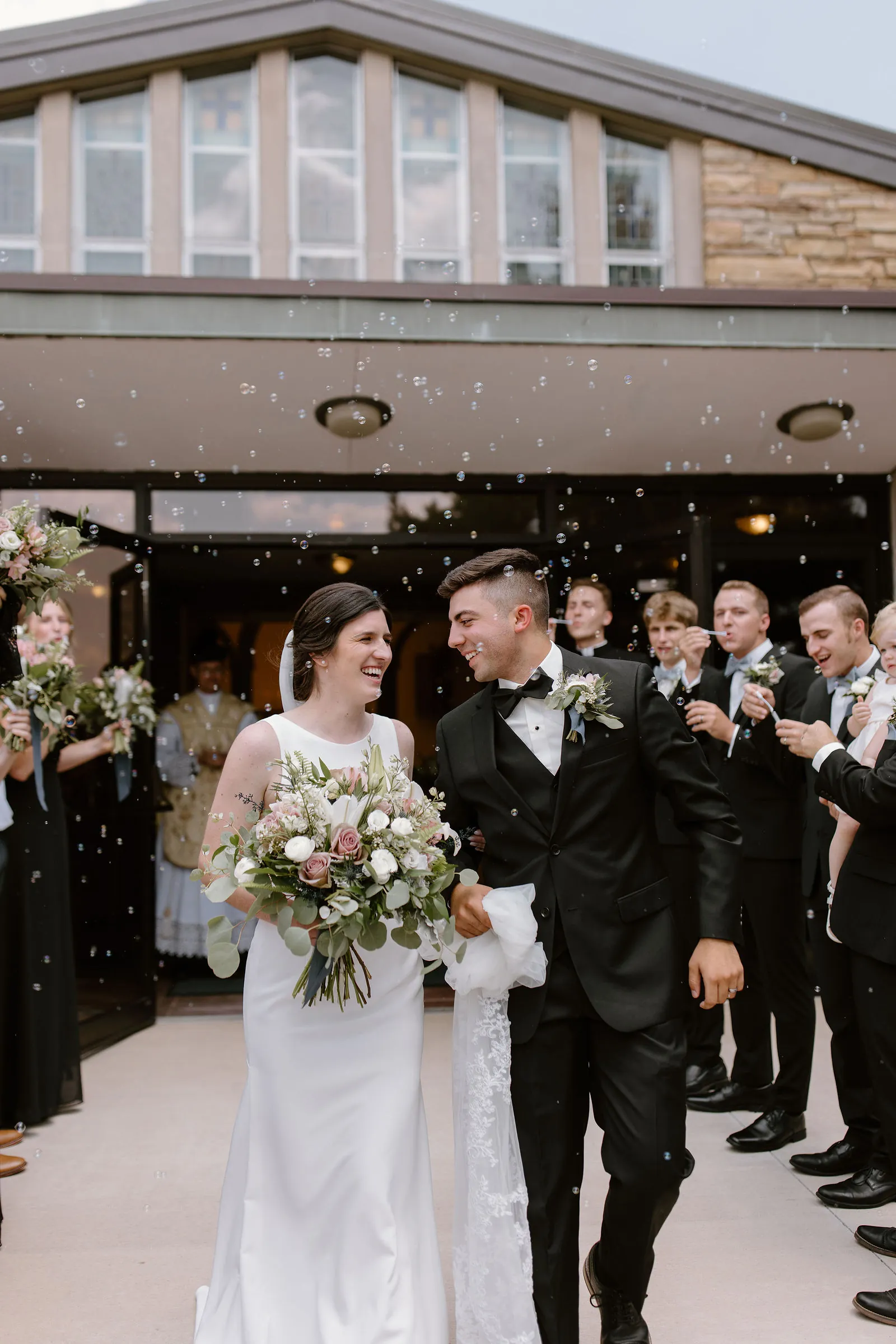 The newly married couple, laughing, walk out of the church through a shower of bubbles, the groomsmen behind them